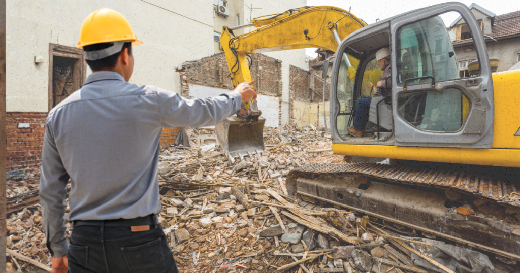 A contractor points at demolition debris as a yellow excavator demolishes a building, highlighting high costs and trade-offs.