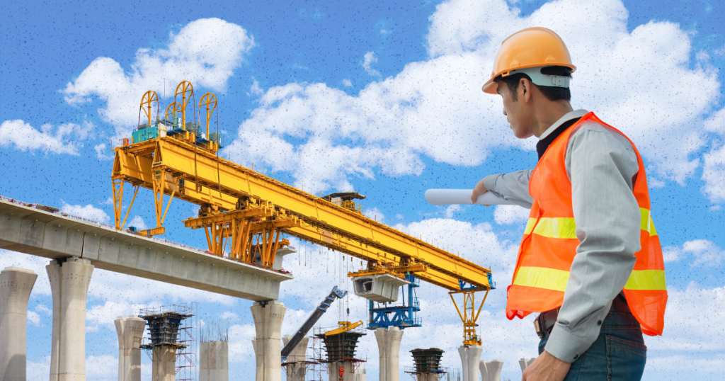 An engineer in a safety vest and hard hat observes a heavy-duty yellow construction crane building an elevated highway or rail.